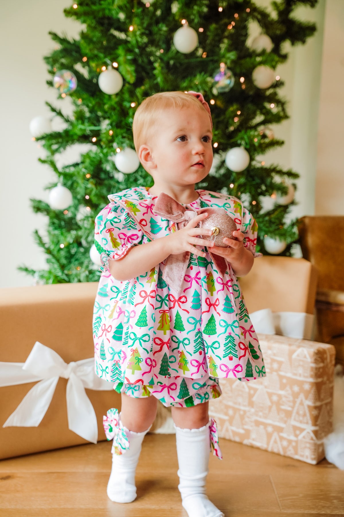Child in a festive dress standing in front of a decorated Christmas tree with presents underneath.