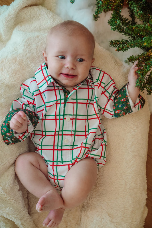 Baby wearing a red and green checkered outfit on a beige blanket with Christmas decorations.