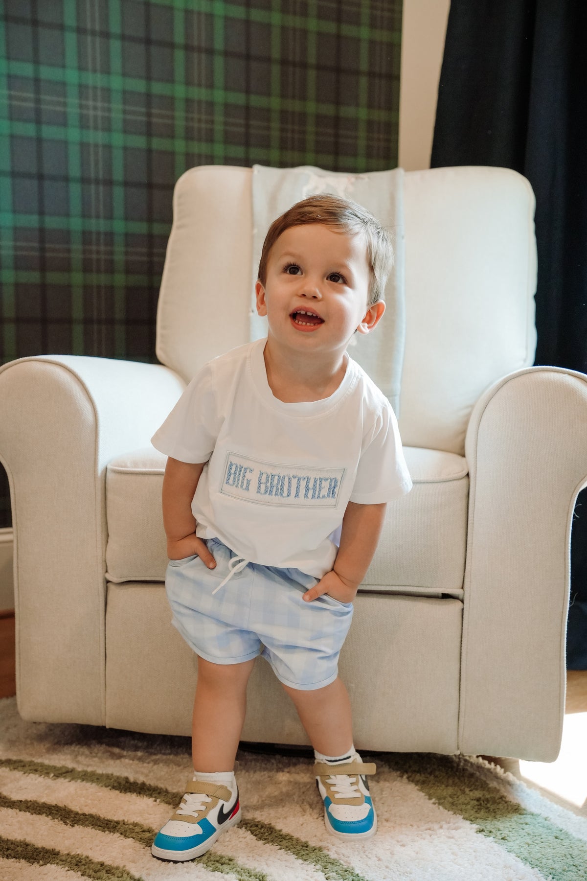 Child wearing a 'Big Brother' shirt standing in front of a white chair.