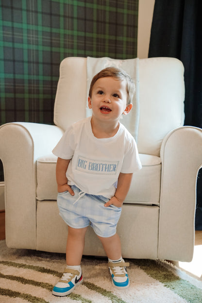 Child wearing a 'Big Brother' shirt standing in front of a white chair.