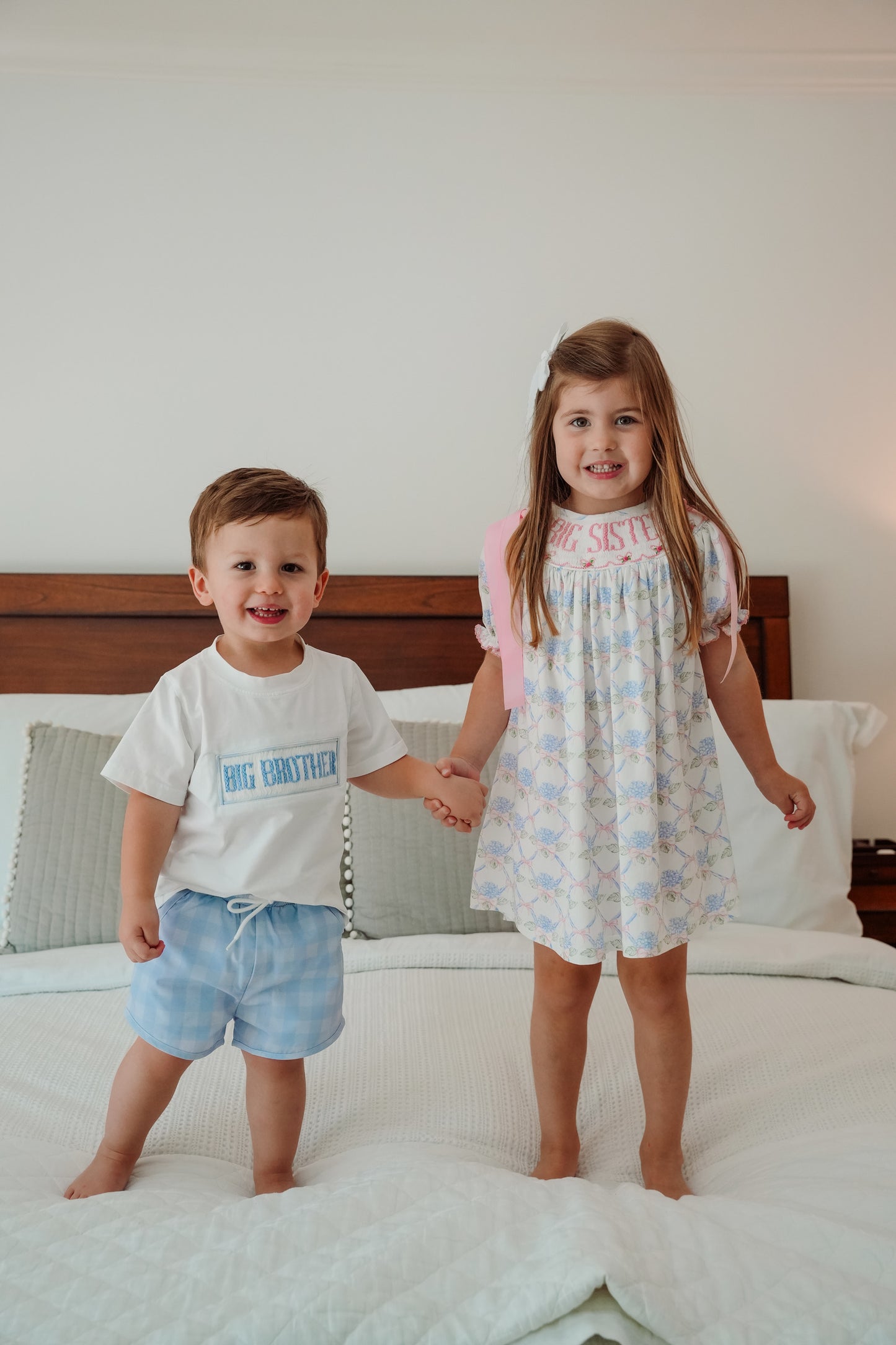 Two children, a big sister and a big brother, standing on a bed holding hands.