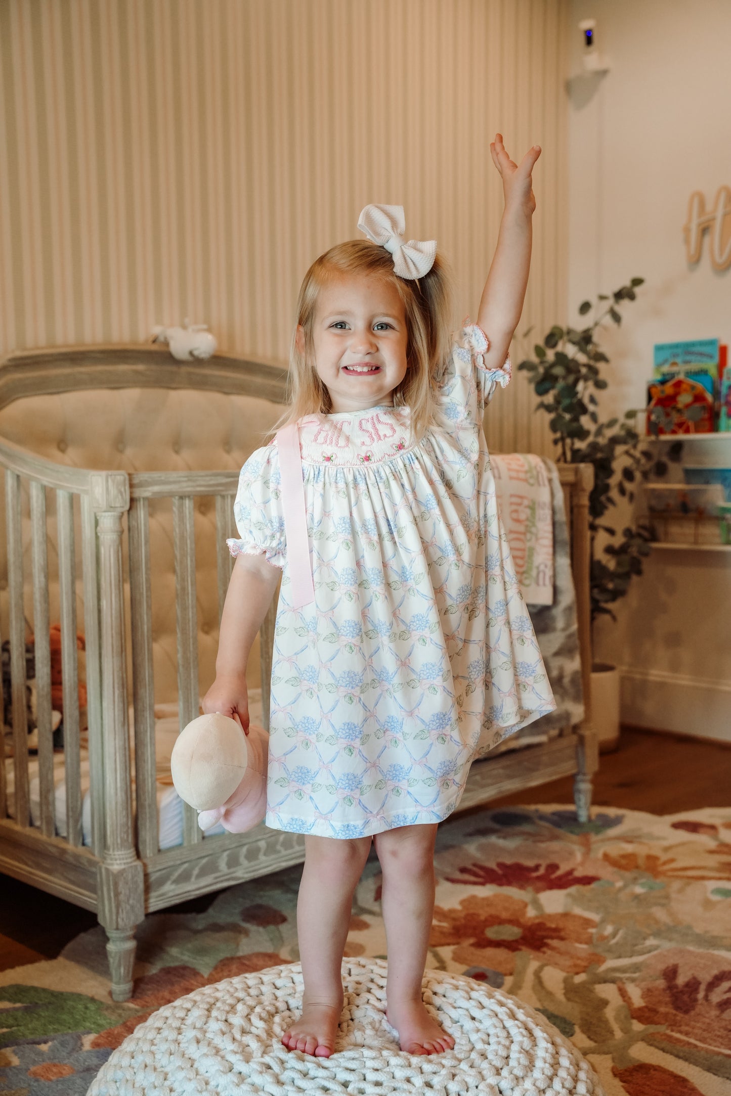 Young girl in a patterned dress standing in a nursery with a crib and decorative rug.