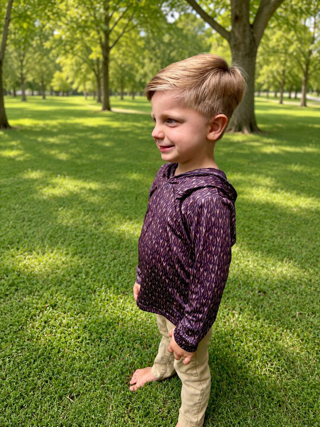 A young boy standing barefoot on a grassy park lawn, wearing a dark purple long-sleeve hooded shirt with a subtle white pattern and khaki pants.