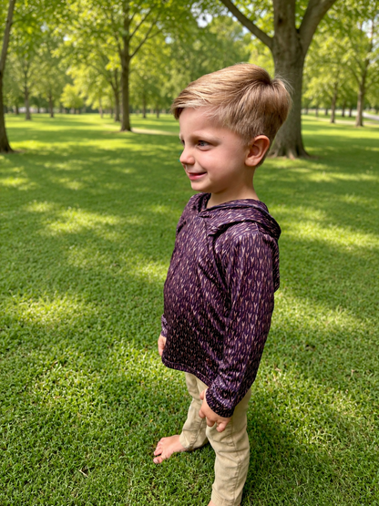 A young boy standing barefoot on a grassy park lawn, wearing a dark purple long-sleeve hooded shirt with a subtle white pattern and khaki pants.