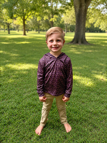 A young boy standing barefoot on a grassy park lawn, wearing a long-sleeve purple patterned hooded shirt and khaki pants.