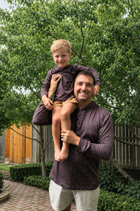A smiling man outdoors wearing a dark patterned long-sleeve shirt and light shorts, carrying a young boy on his shoulders. The boy is wearing a matching dark patterned long-sleeve shirt and tan shorts.
