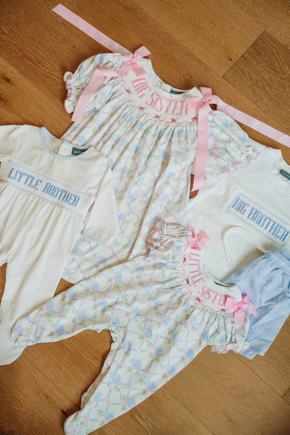 Three baby clothing sets laid out on a wooden floor: a cream long-sleeve top with blue 'LITTLE BROTHER' text, a white smocked dress with pink 'BIG BROTHER' text and pink ribbon bows, and a cream floral romper with pink 'BIG BROTHER' text and pink ribbon bows, plus light blue shorts.