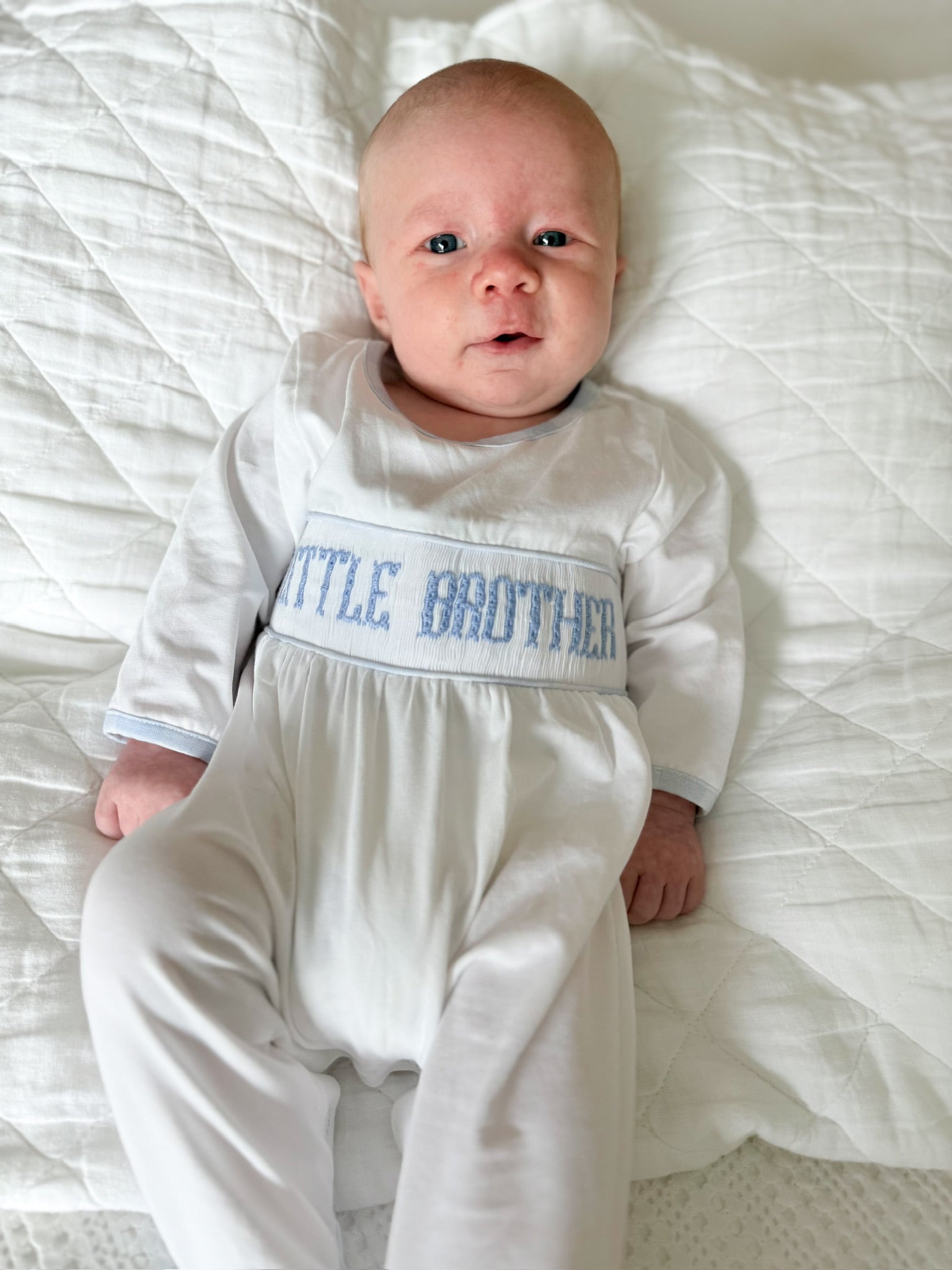 Newborn baby wearing Sawyer + Spade Little Brother Smocked Footie with blue hand-smocked embroidery lying on a white quilt