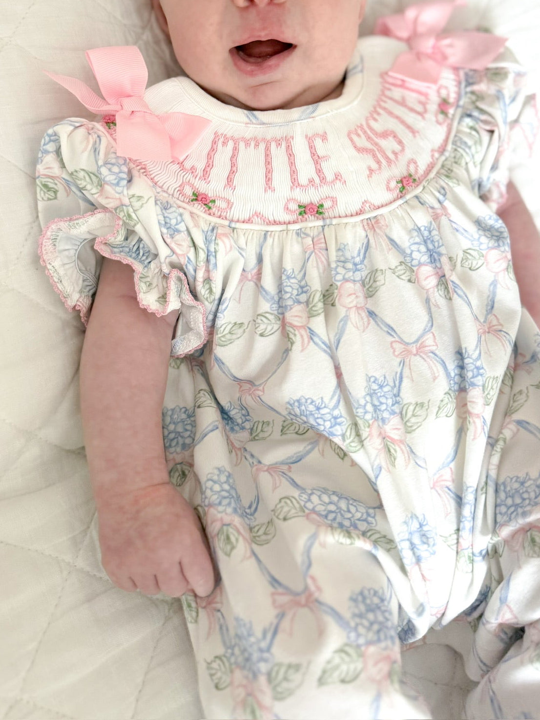 Close-up of Sawyer + Spade Little Sister smocked newborn footie featuring hand-smocked "Little Sister" embroidery, pink bow accents, and a pastel floral print