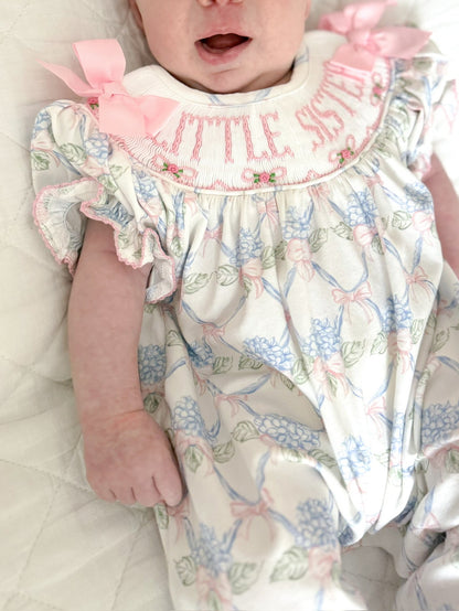 Close-up of Sawyer + Spade Little Sister smocked newborn footie featuring hand-smocked "Little Sister" embroidery, pink bow accents, and a pastel floral print
