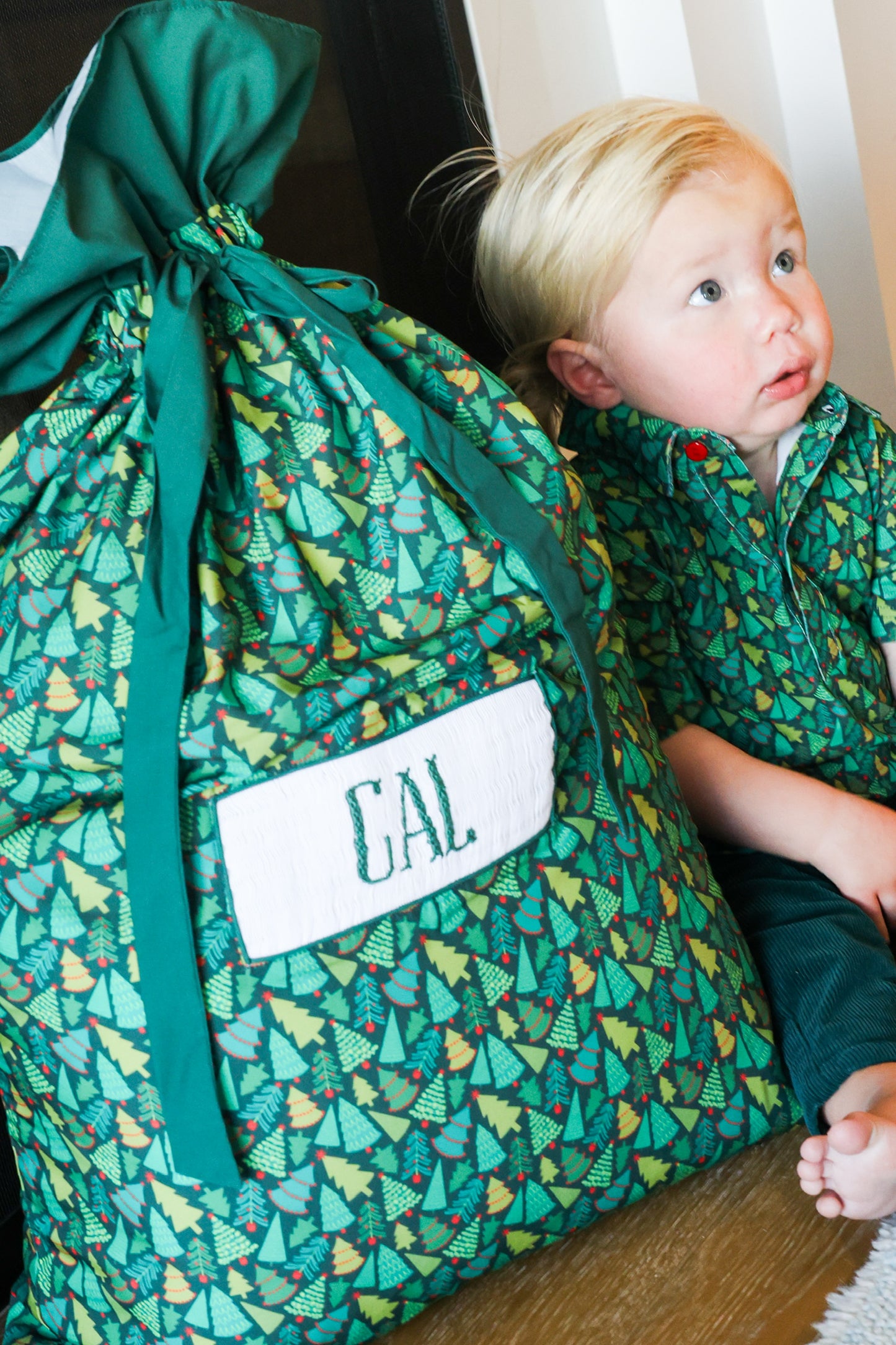 A child wearing a matching green patterned shirt sits beside a large green drawstring bag with a Christmas tree print and a white patch reading 'GAL'.