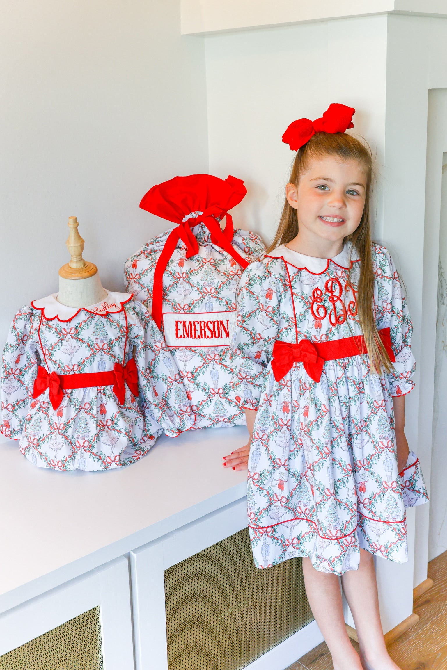 A young girl stands indoors wearing a white dress with a red and gray toile pattern and a large red bow, next to two matching dresses displayed on mannequins. One dress has a name patch reading EMERSON.