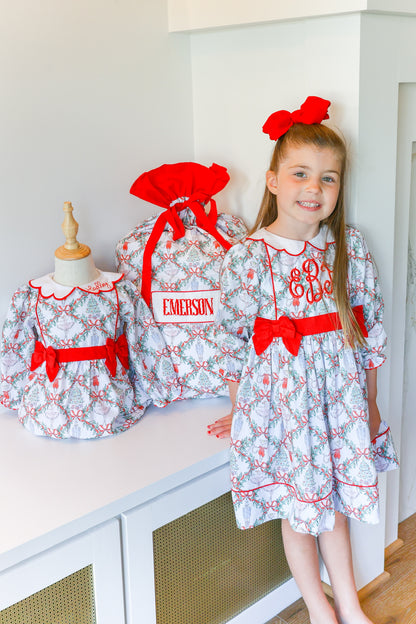 A young girl stands indoors wearing a white dress with a red and gray toile pattern and a large red bow, next to two matching dresses displayed on mannequins. One dress has a name patch reading EMERSON.