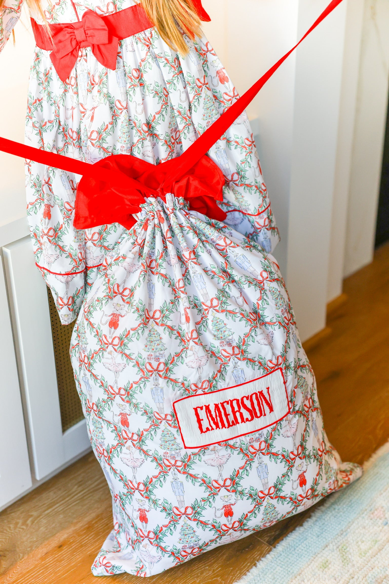 A Christmas-themed drawstring gift bag with a red ribbon and a personalized name patch reading 'EMERSON', shown on a wooden floor next to a child wearing a matching printed dress.