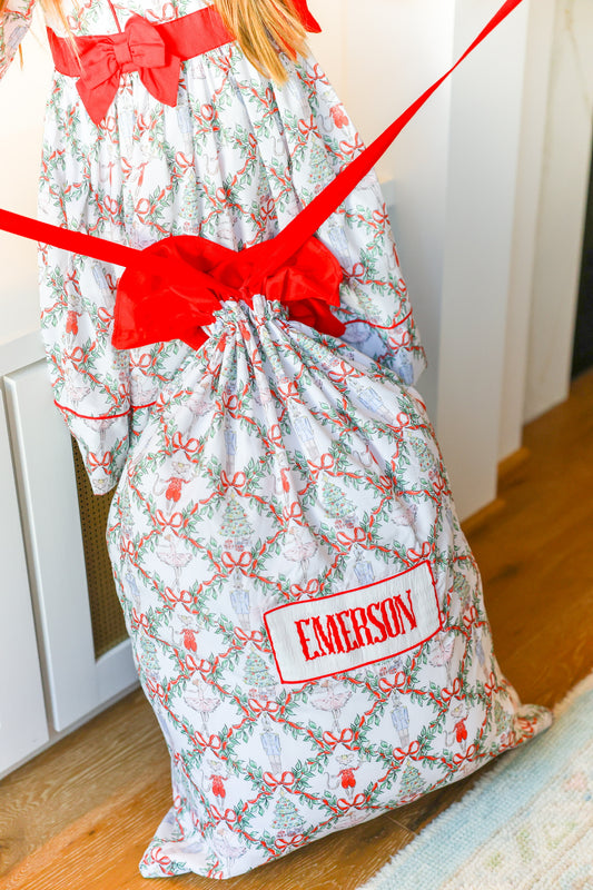 A Christmas-themed drawstring gift bag with a red ribbon and a personalized name patch reading 'EMERSON', shown on a wooden floor next to a child wearing a matching printed dress.