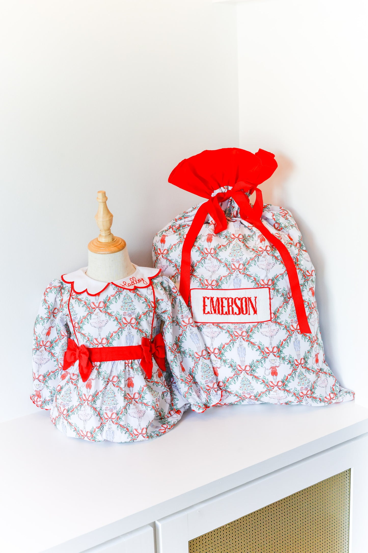 A small dress displayed on a mannequin next to a larger fabric gift bag with a red drawstring closure and a personalized name patch reading EMERSON. The fabric has a repeating holiday pattern in red, green, and white.