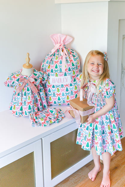 A young girl stands barefoot in a bright room next to a white cabinet, holding a gift box while wearing a colorful holiday-themed dress. On the cabinet are matching dress bags and a smaller dress displayed on a mannequin, all in a coordinated print with bows and trees.