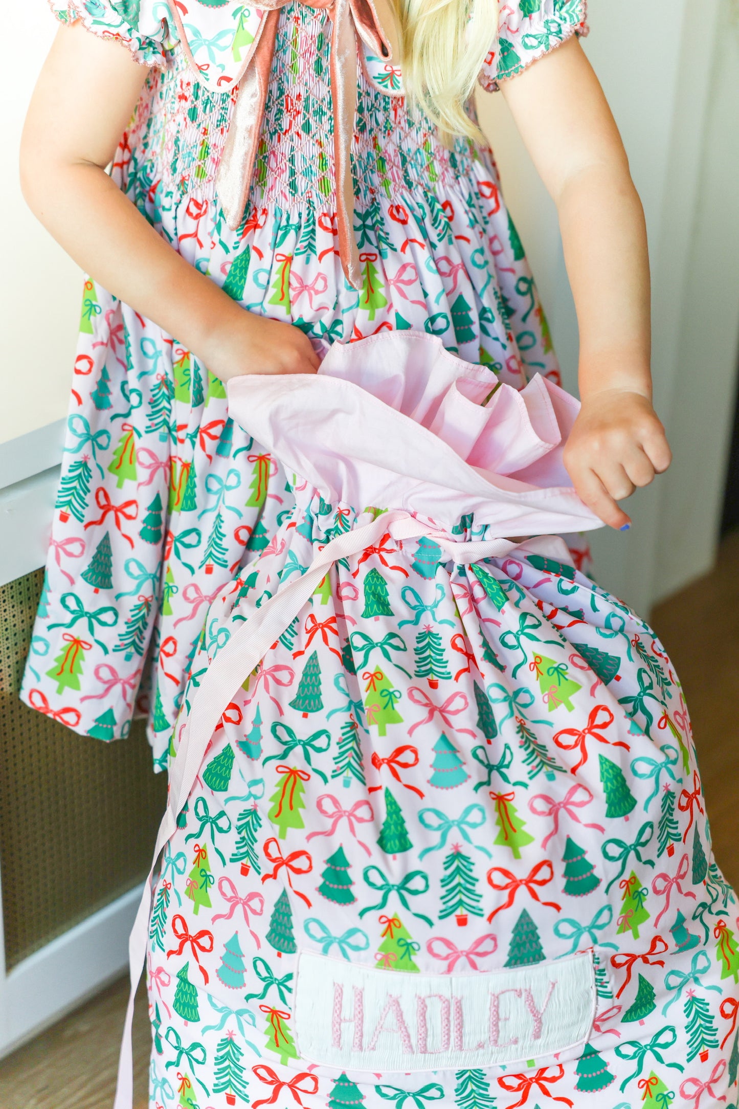 A child wearing a festive Christmas-themed dress with a matching gift bag printed with red bows, green trees, and pink ribbons. The bag is personalized with the name 'HADLEY' on a white patch.