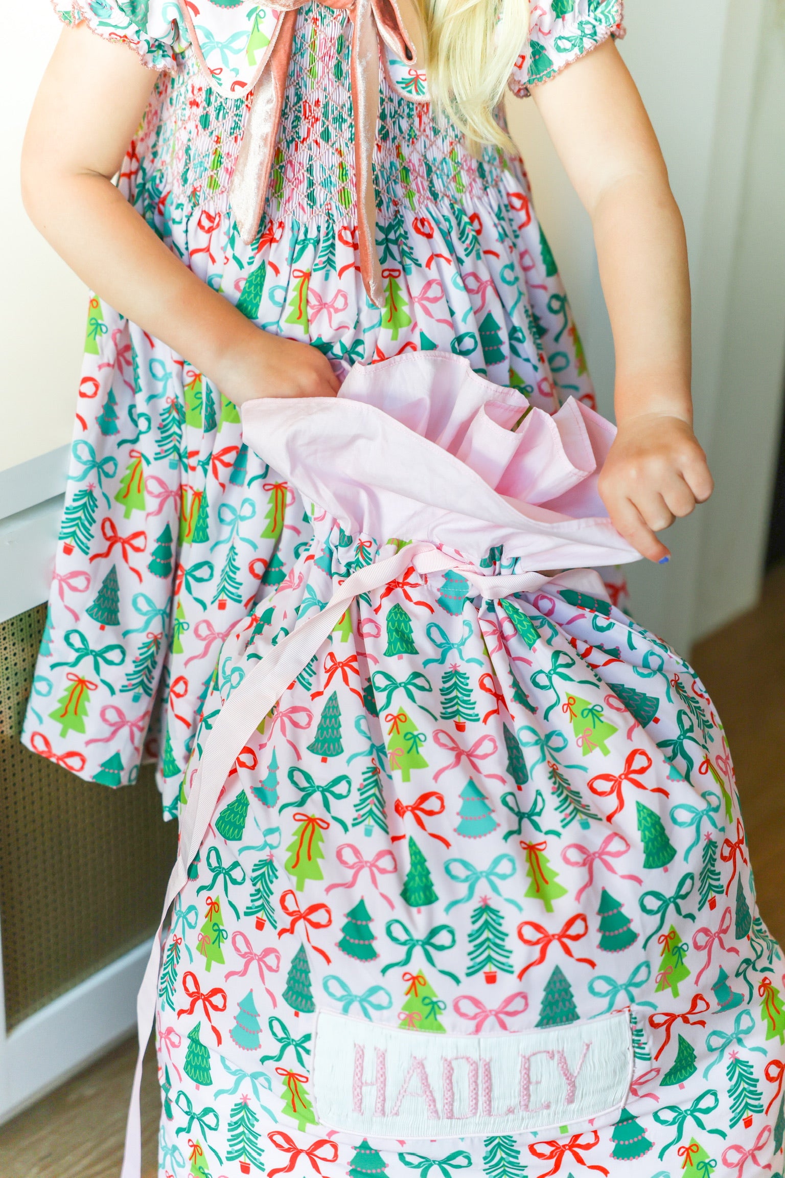 A child wearing a festive Christmas-themed dress with a matching gift bag printed with red bows, green trees, and pink ribbons. The bag is personalized with the name 'HADLEY' on a white patch.