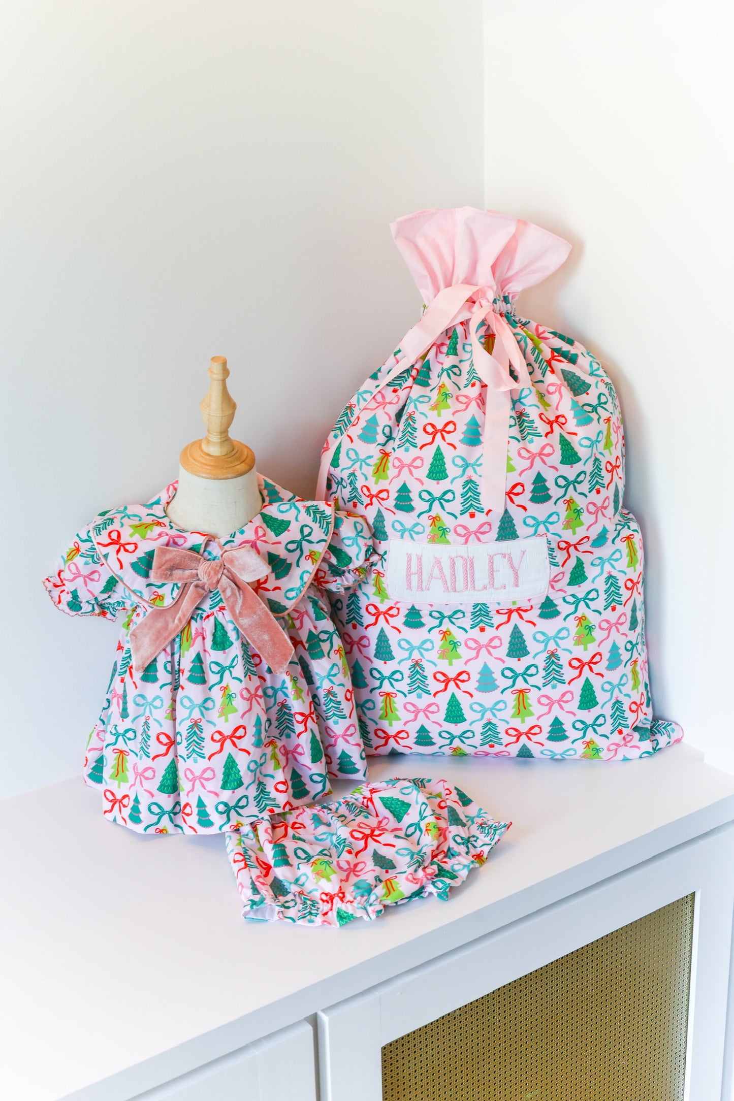 A matching baby and doll clothing set with a colorful Christmas tree and bow pattern, featuring a larger dress on a mannequin, a smaller dress, a matching ruffled headband, and a pink drawstring bag labeled 'HANLEY'.