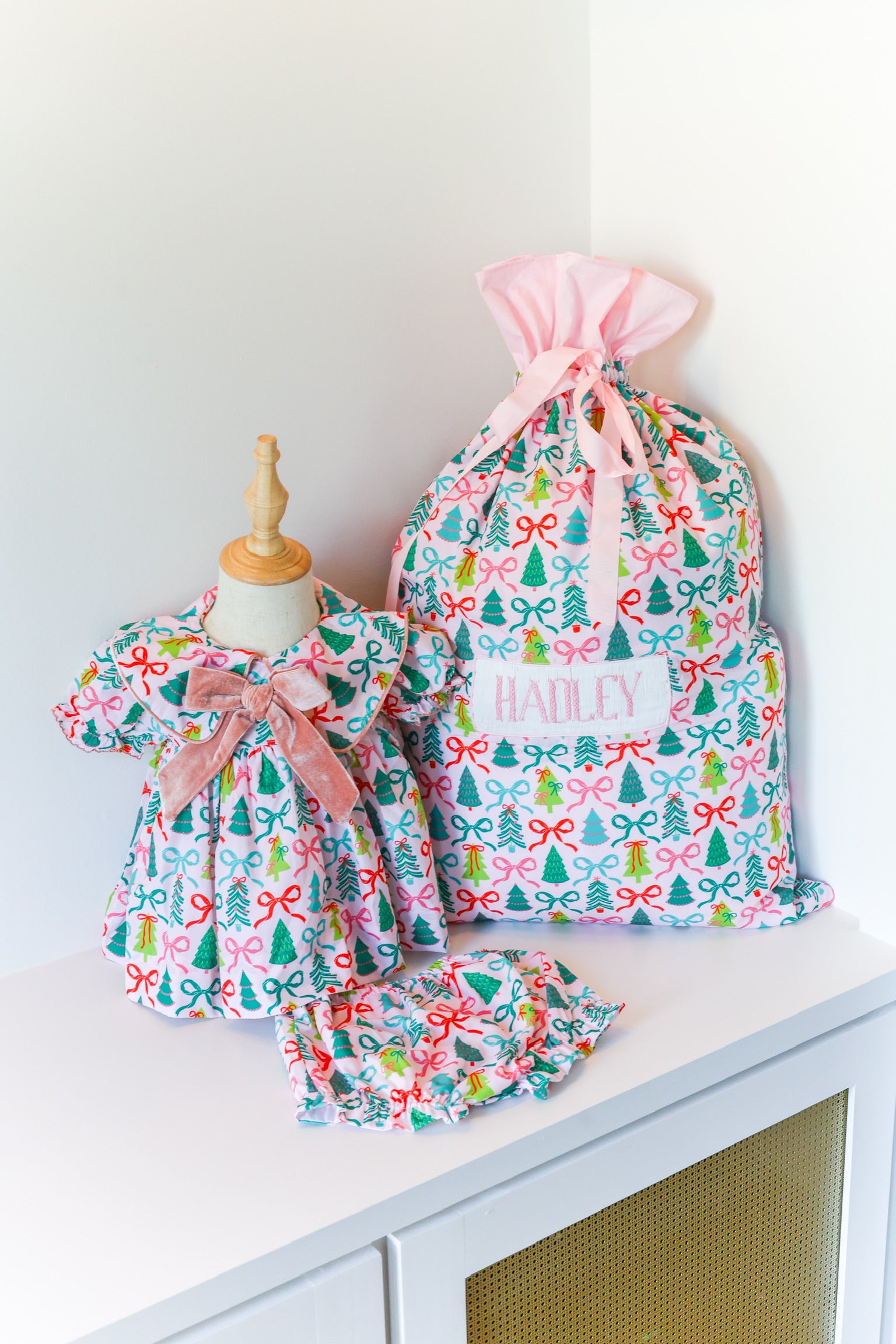 A matching baby and doll clothing set with a colorful Christmas tree and bow pattern, featuring a larger dress on a mannequin, a smaller dress, a matching ruffled headband, and a pink drawstring bag labeled 'HANLEY'.