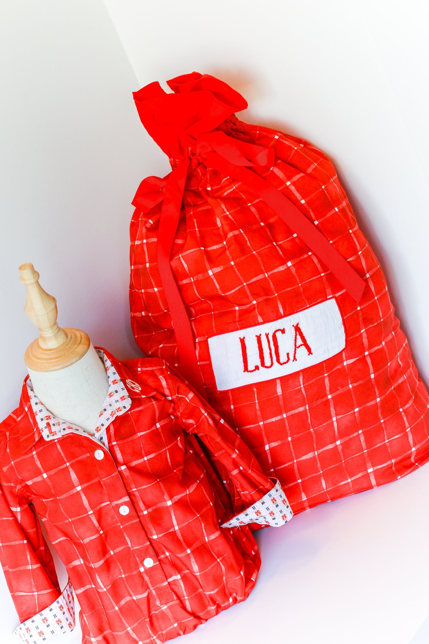 A red and white plaid baby gift set featuring a personalized red drawstring bag with a white patch reading 'LUCA' and a matching red plaid baby pajama set displayed on a small mannequin.