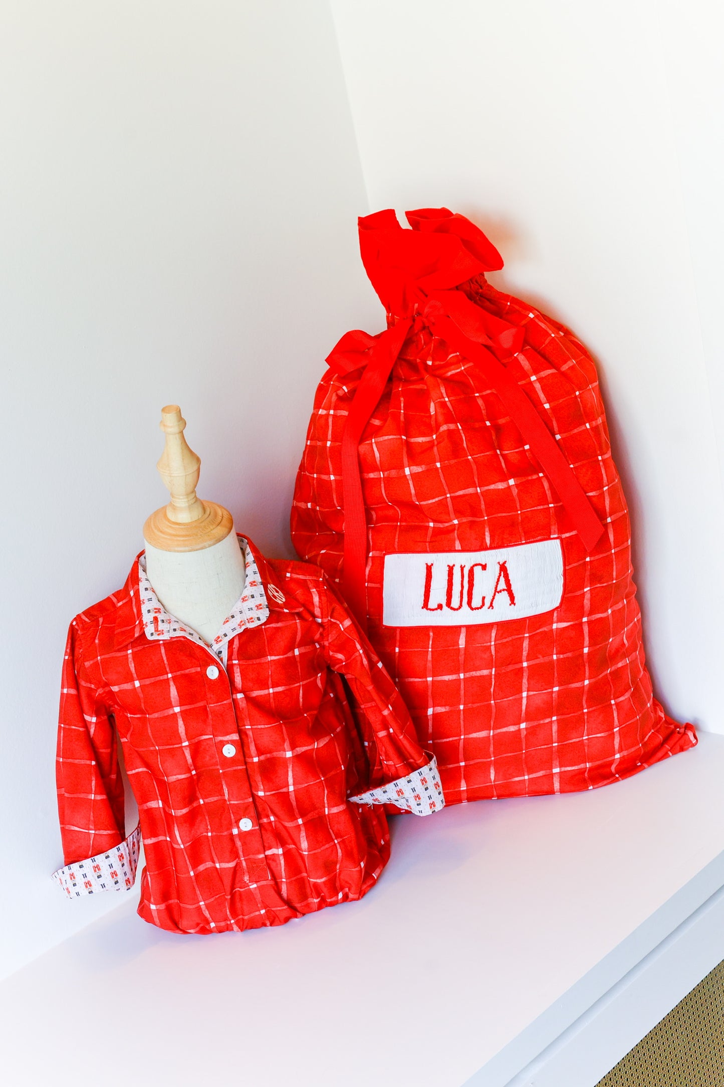 A red and white plaid children's pajama set displayed on a small mannequin next to a matching drawstring bag with the name 'LUCA' embroidered on a white patch.